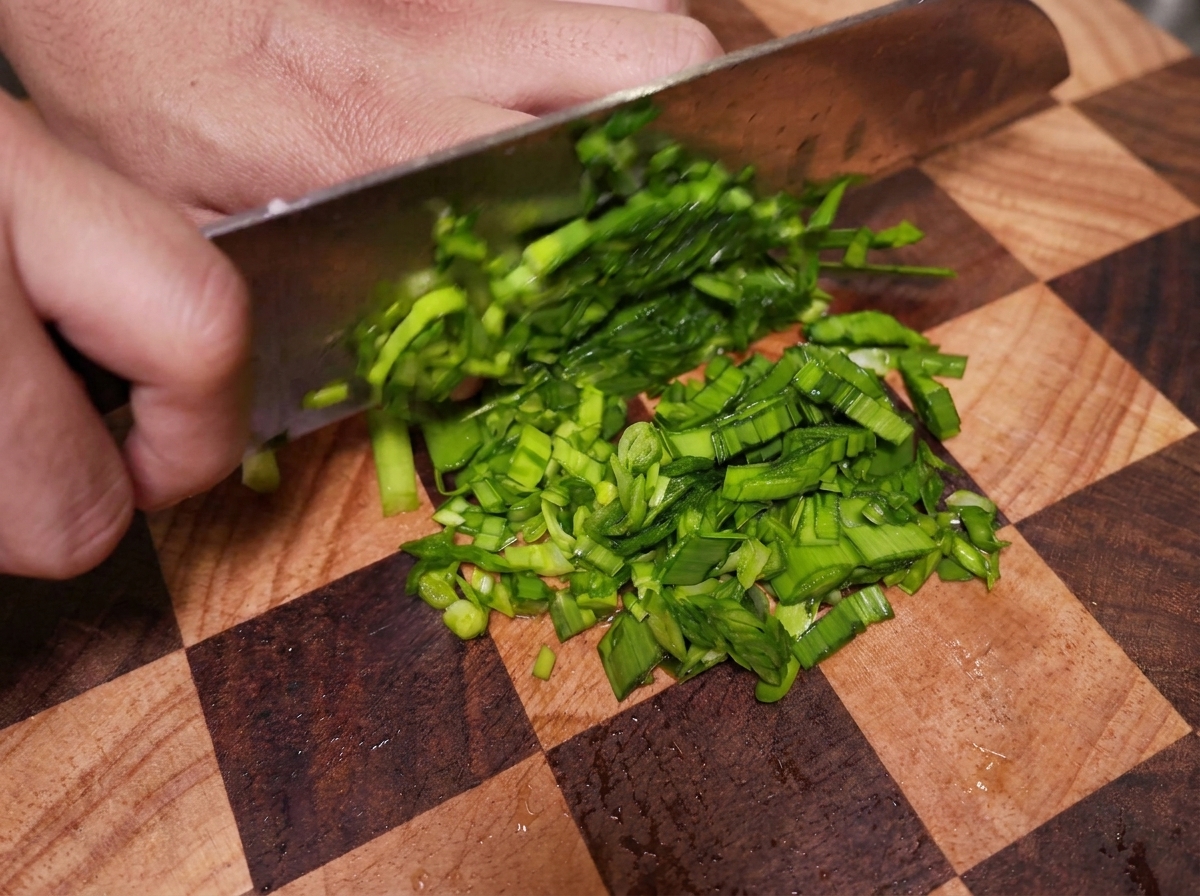 A person using a cleaver to slice bright green chives into fine pieces on a checkered wooden cutting board.