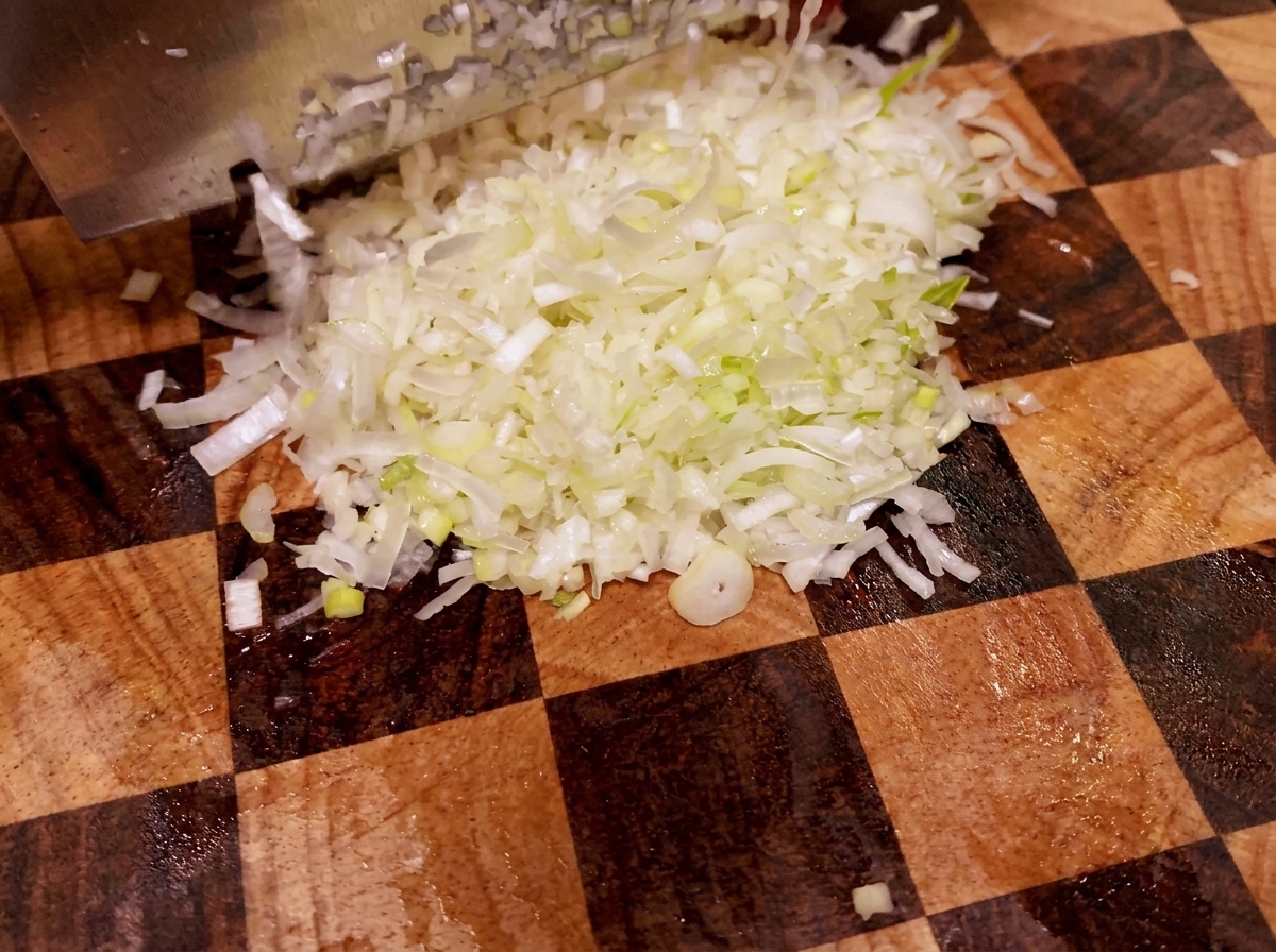 A sharp knife chopping a white green onion stalk on a wooden cutting board.