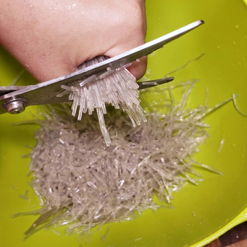 Kitchen scissors cutting through a bundle of soaked, transparent glass noodles inside a green mixing bowl.