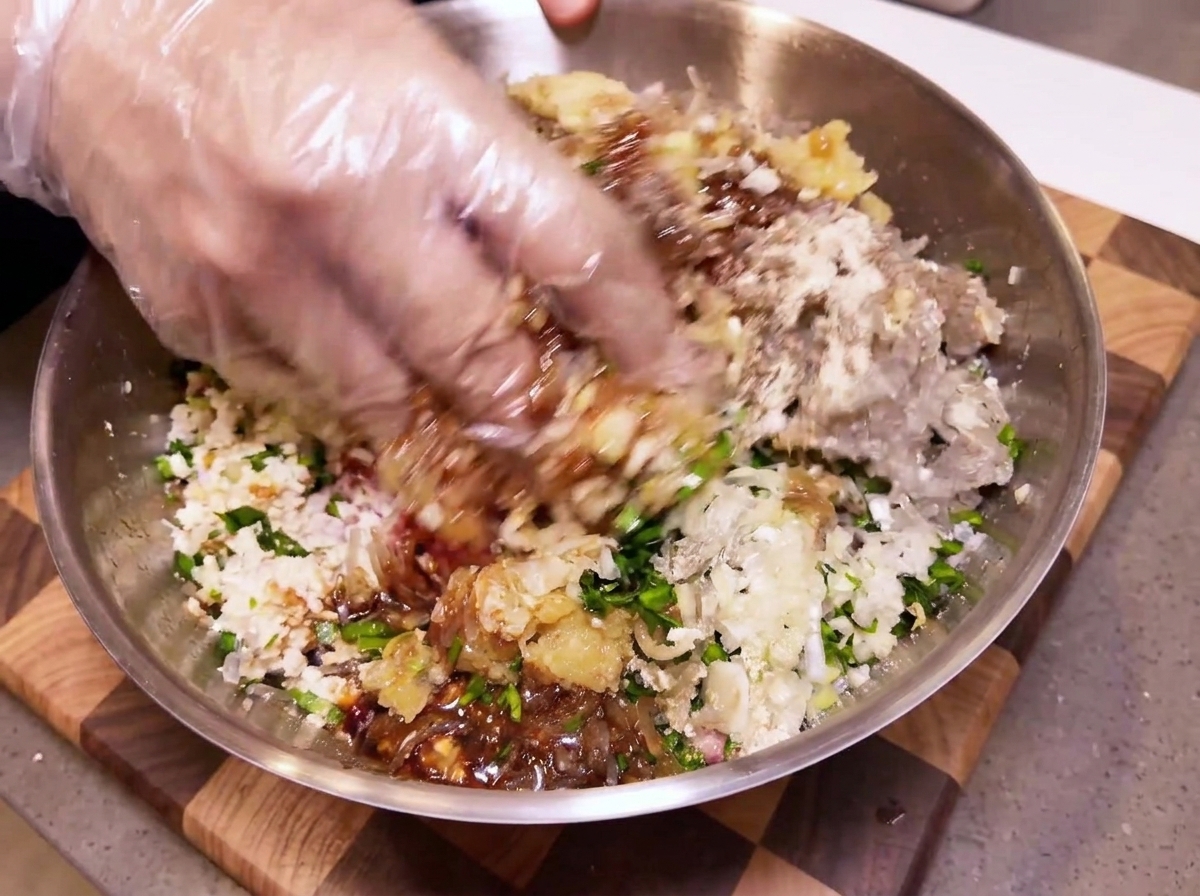 A hand in a clear plastic glove mixing meat and vegetables inside a large metal bowl.
