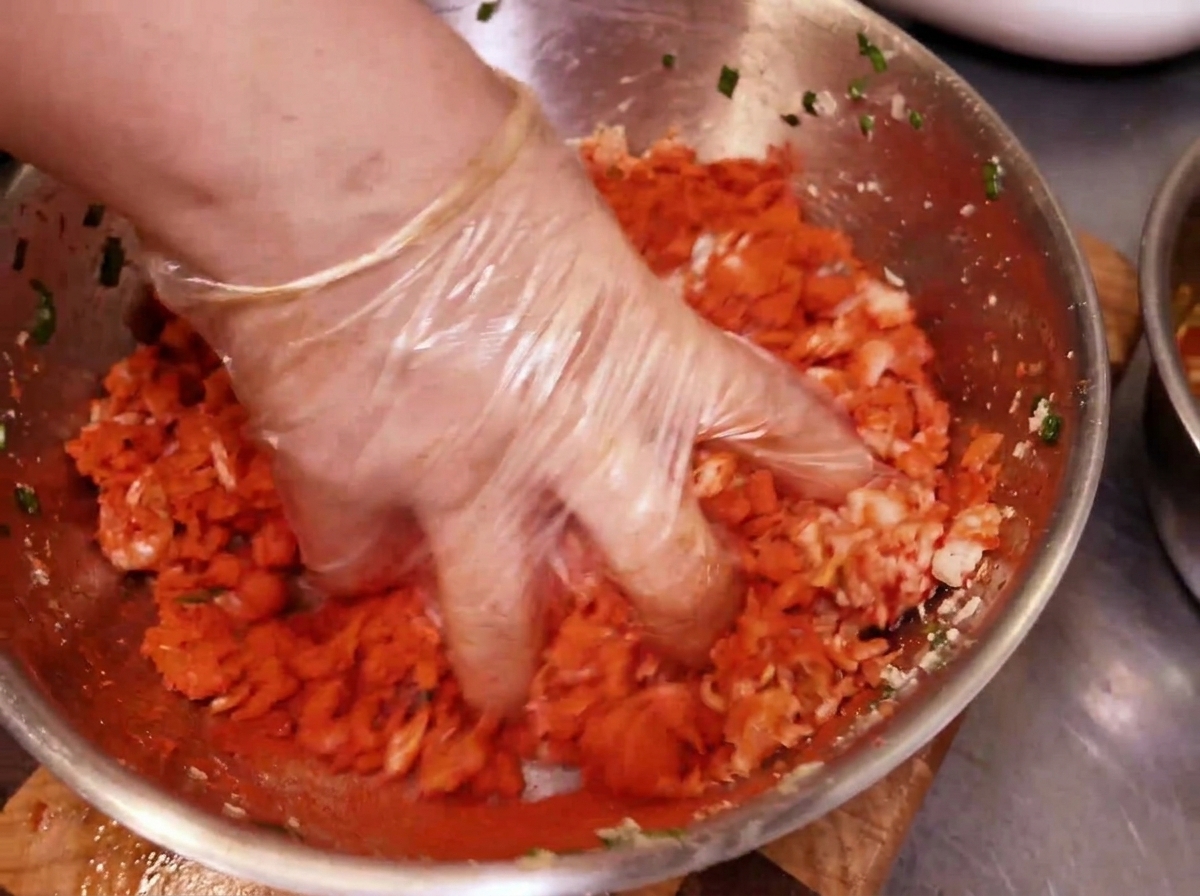A gloved hand kneading and mixing a vibrant red kimchi and pork filling inside a metal bowl.