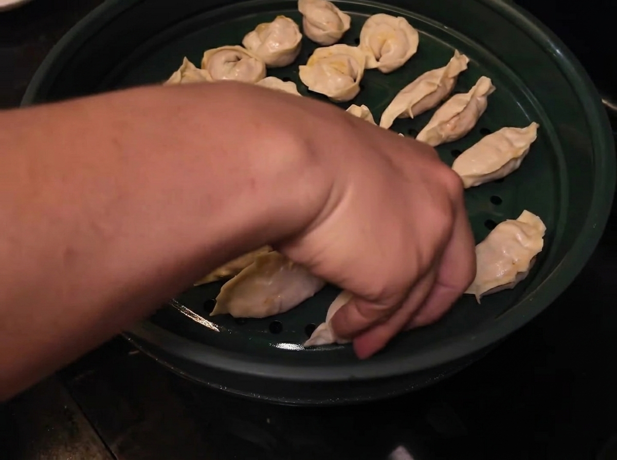 A hand arranging neatly folded raw dumplings into a green steamer basket lined with holes.