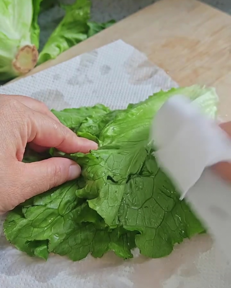 Hands drying fresh, green lettuce leaves using white paper towels on a wooden board.