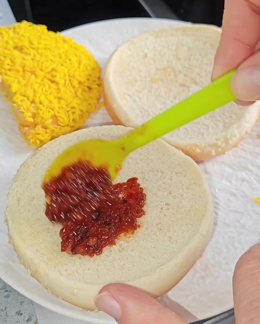 A small spatula spreading red chili sauce onto the inner face of a toasted burger bun.