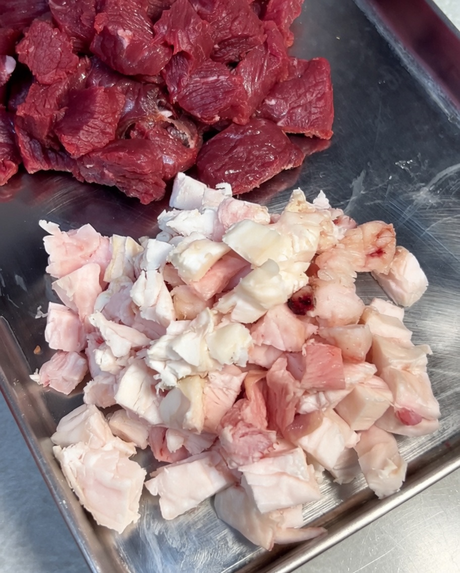 Beef and fat pieces being seasoned with salt and black pepper in a stainless steel bowl.