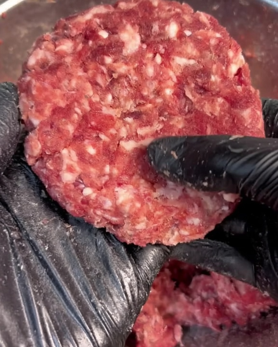 Hand in a black glove shaping seasoned ground beef into a round patty.