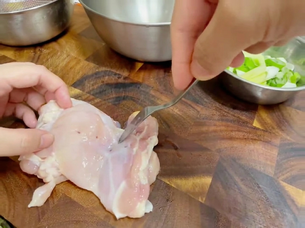 Close-up of a fork piercing a raw deboned chicken thigh on a wooden cutting board.
