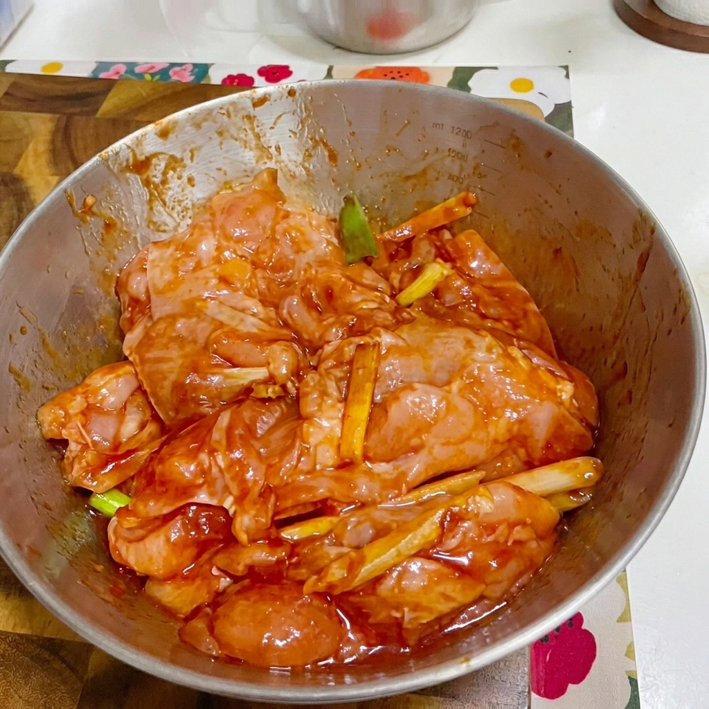 A metal bowl of marinated chicken covered with plastic wrap.