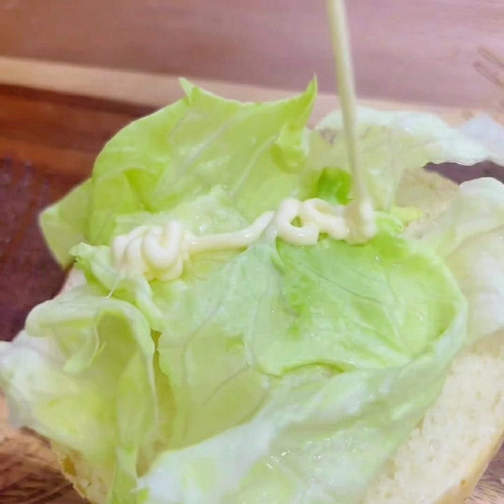 White mayonnaise being squeezed from above in a zigzag line onto the fresh green lettuce resting on the burger bun.