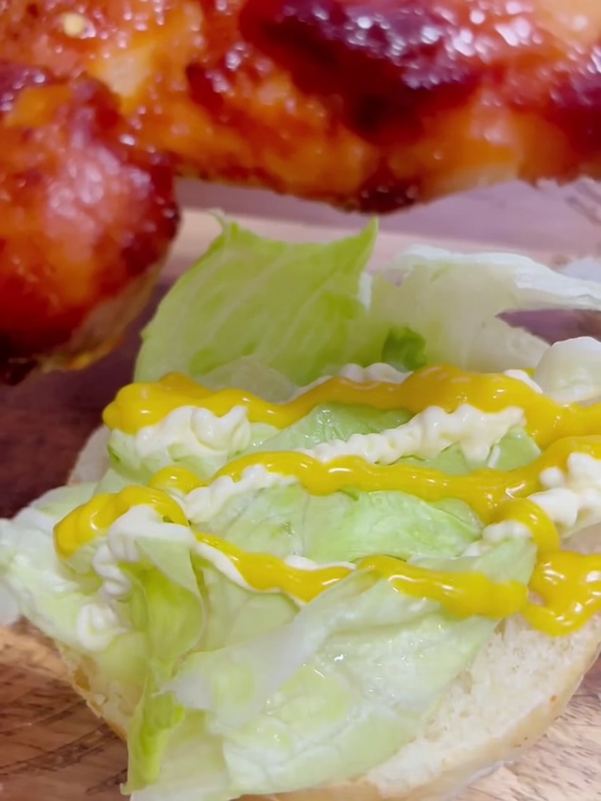 A piece of glazed teriyaki chicken thigh being placed over green lettuce dressed with yellow honey mustard and white mayonnaise.
