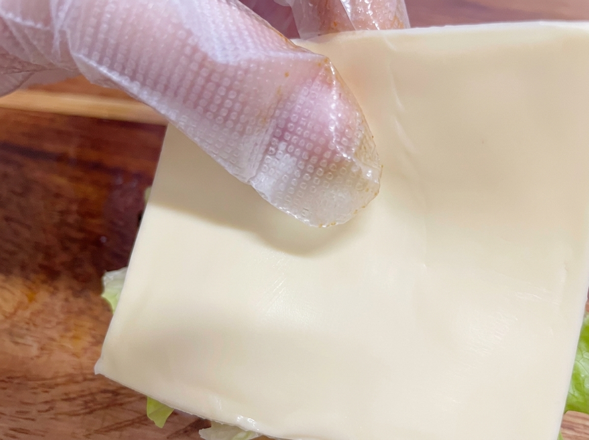 A hand wearing a plastic glove carefully placing a square slice of white cheese onto the burger assembly.