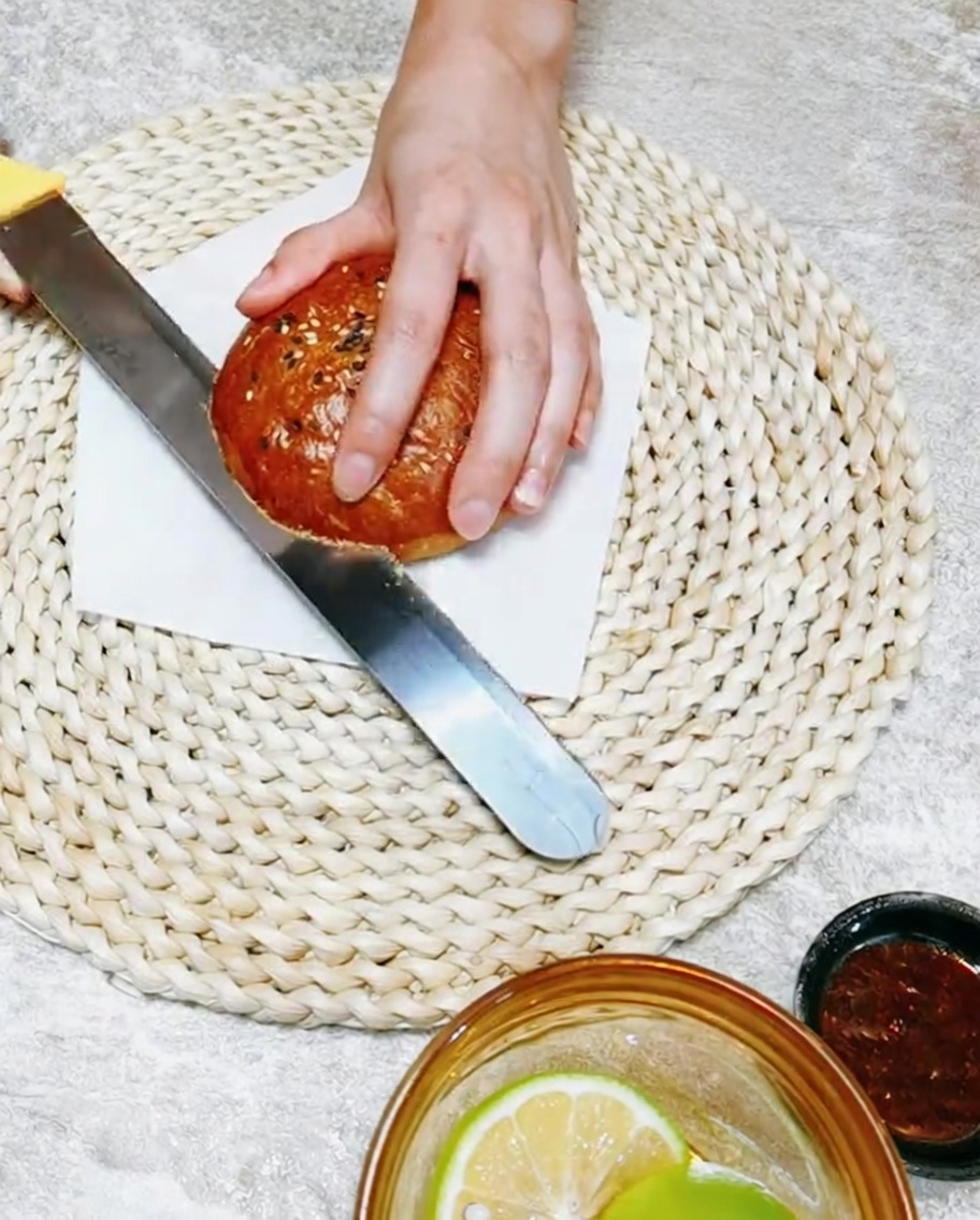A hand holding a knife while slicing a soft, handmade burger bun in half on a wooden cutting board.