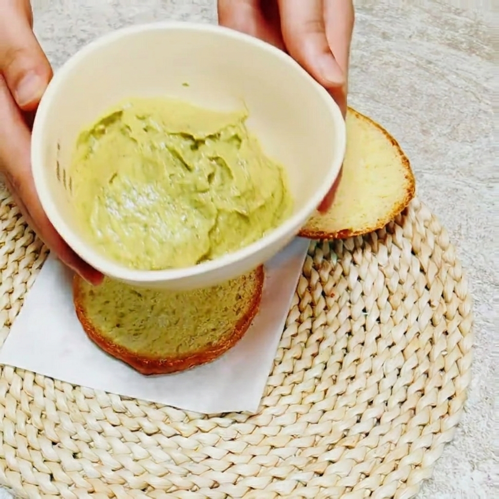 Close-up of a knife spreading bright green avocado basil sauce onto the interior of a burger bun.