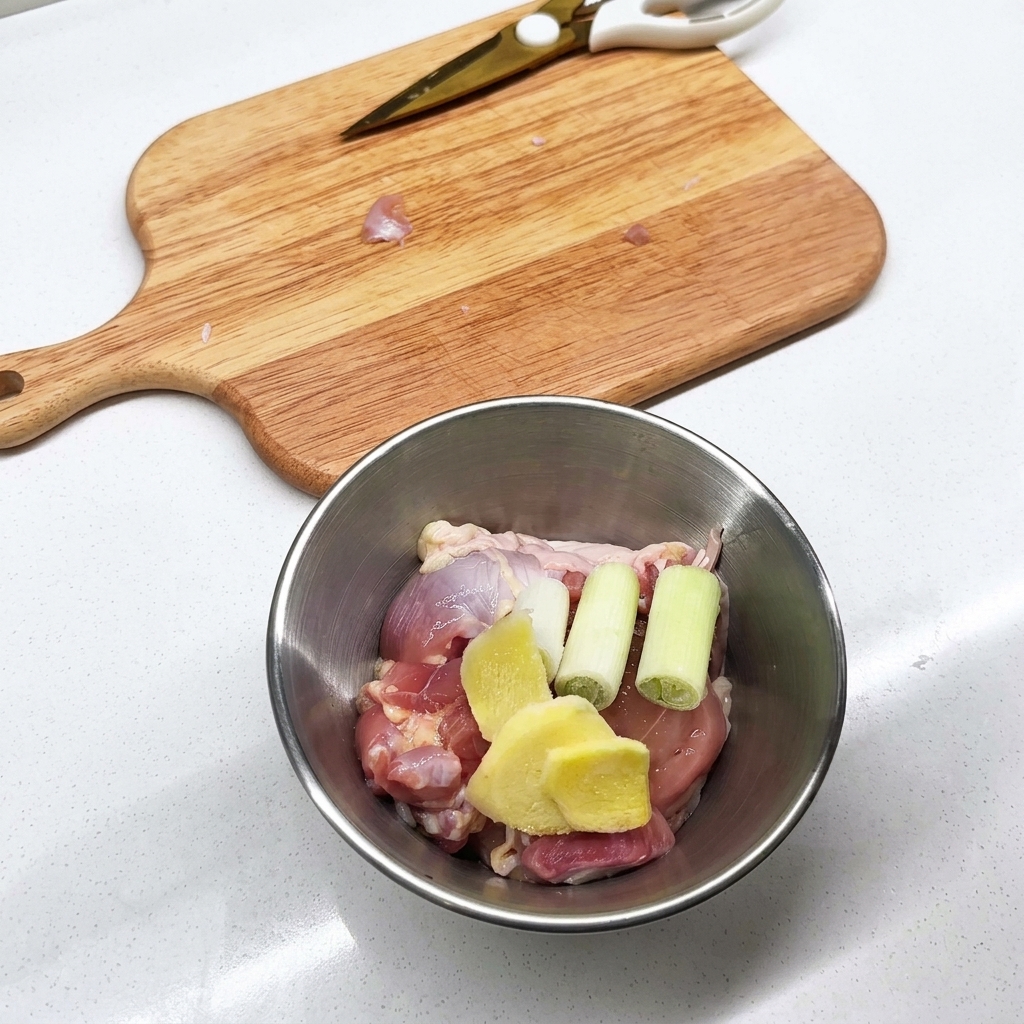 Metal bowl containing raw chicken leg pieces, sliced ginger, and green onion stalks.