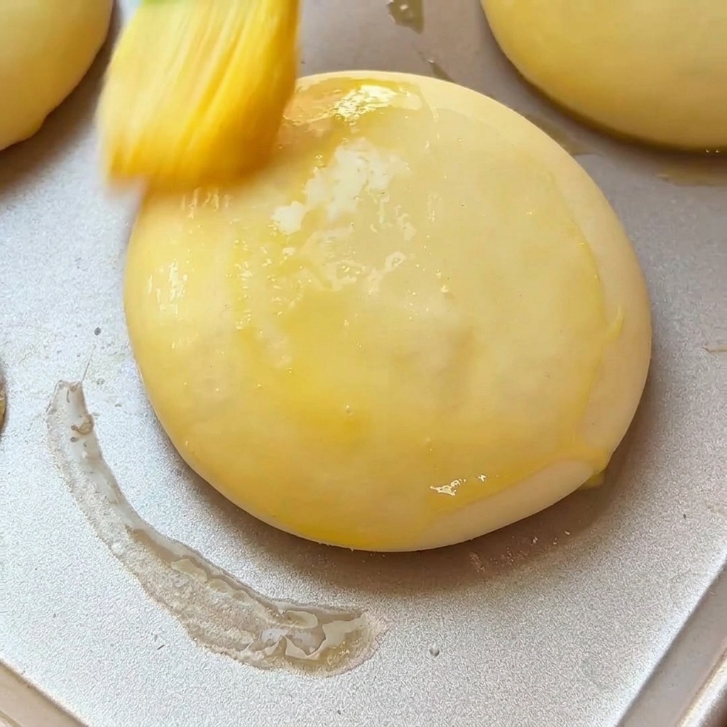 A pastry brush applying egg wash to a soft, proofed burger bun sitting on a metal baking tray.