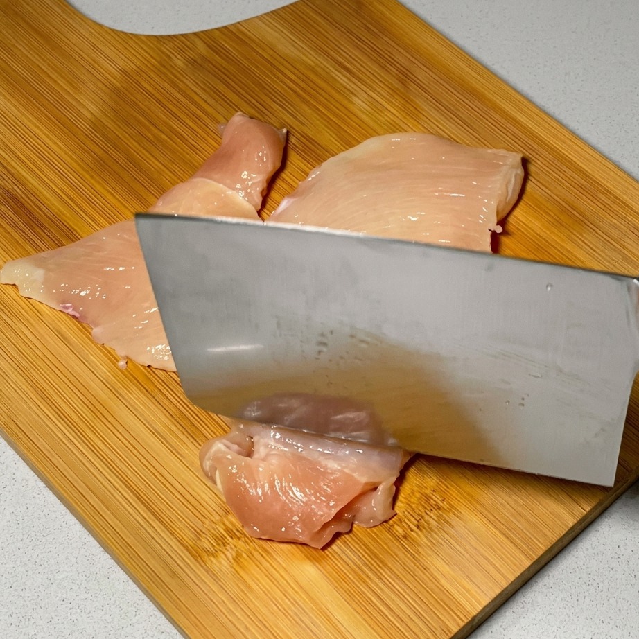 A kitchen knife being used to tenderize raw chicken slices on a wooden surface.