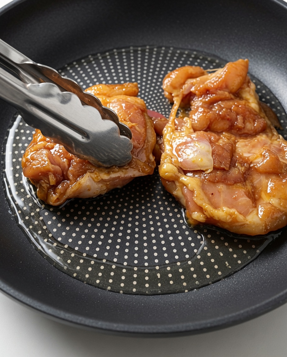 Raw, marinated chicken thighs being laid into a hot, oiled skillet using metal tongs.