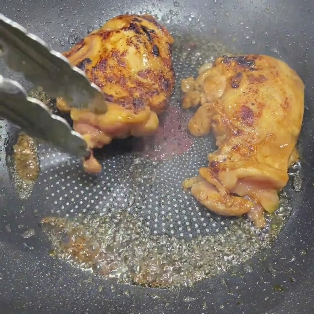 Golden-brown teriyaki glazed chicken thighs being turned over in a skillet with kitchen tongs.