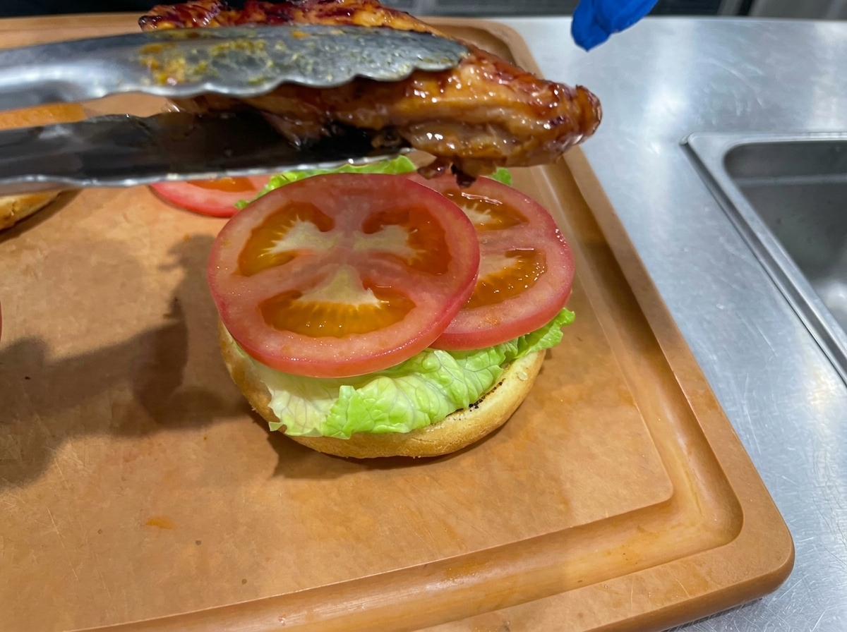 A cooked teriyaki chicken thigh being placed with tongs onto a burger bun layered with lettuce and tomato slices.