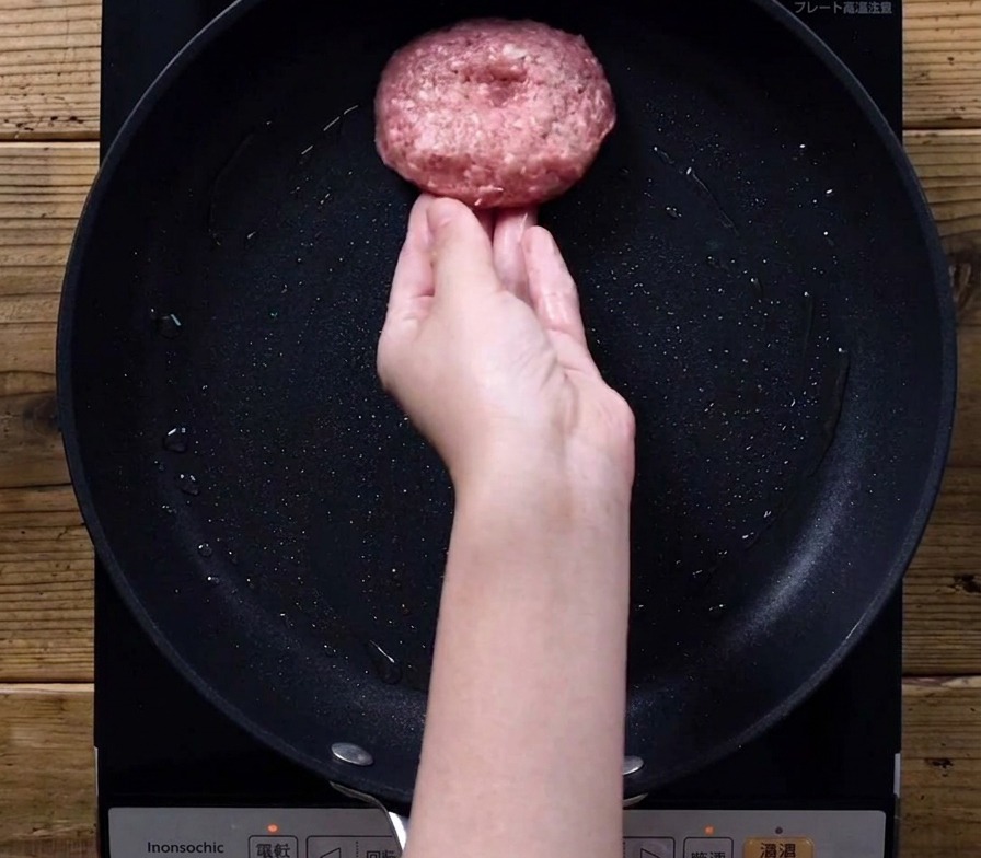 A hand gently placing a thick, round raw meat patty into a black frying pan on an induction stove.