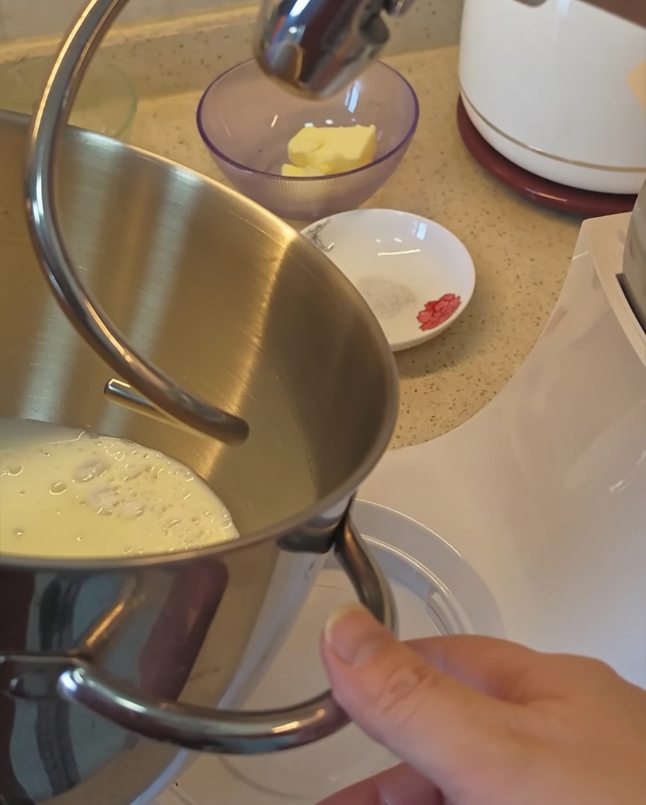 A close-up view of the stand mixer bowl containing the dry ingredients and milk for the bun dough.