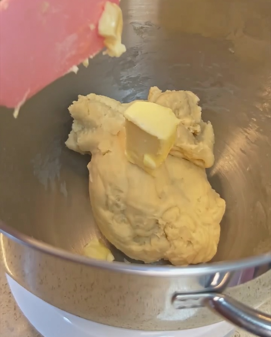 A knob of butter being added to the partially kneaded dough inside the stainless steel mixing bowl.