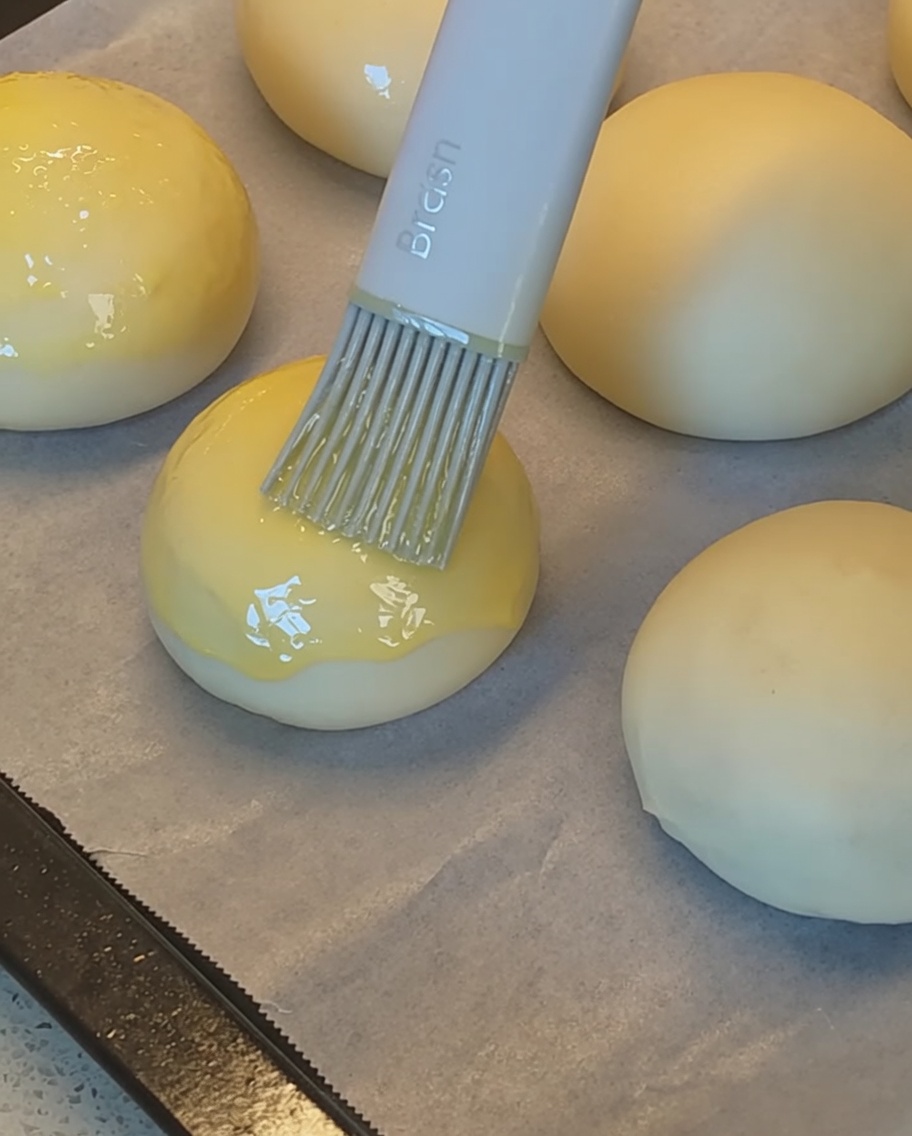 A silicone brush applying egg wash to the tops of shaped bun dough pieces before they are sprinkled with sesame seeds.