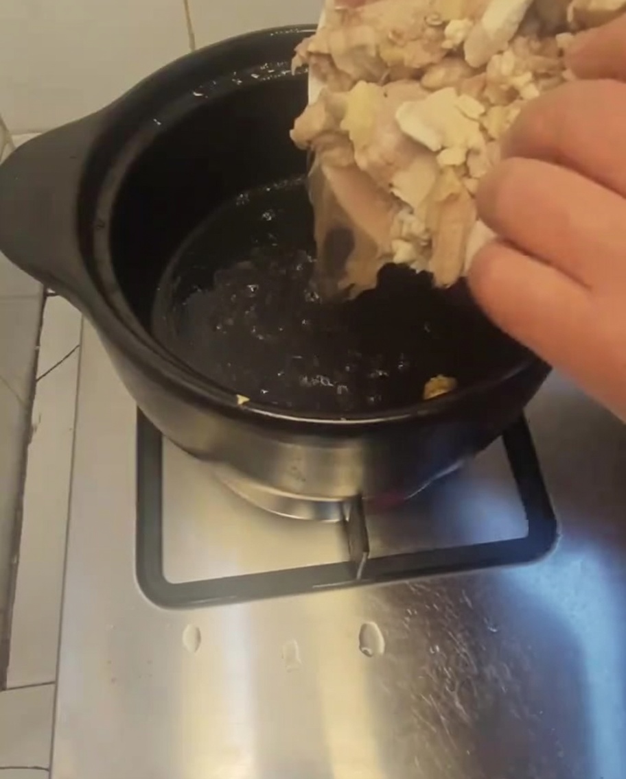 Blanched chicken pieces being poured from a white plate into a dark, preheated clay pot.