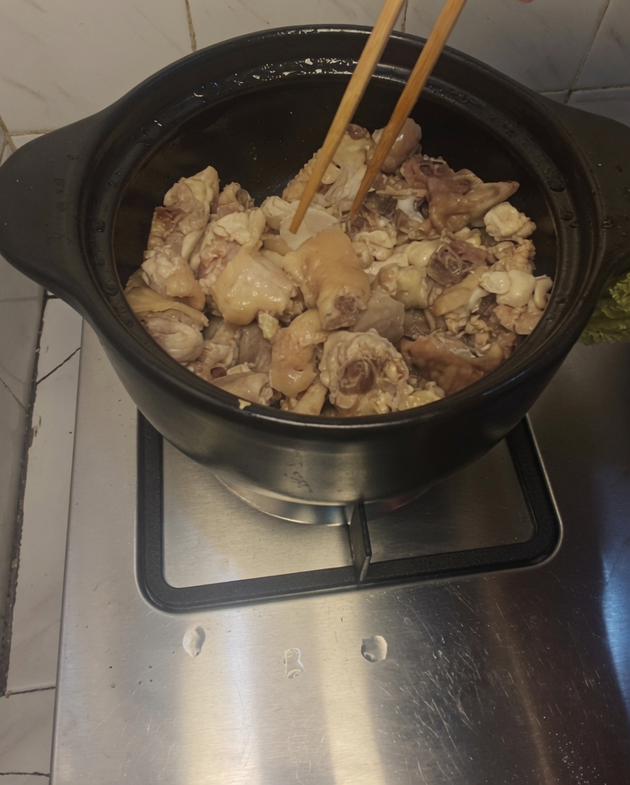 Wooden chopsticks stirring chunks of chicken and ginger slices inside a heated clay pot.