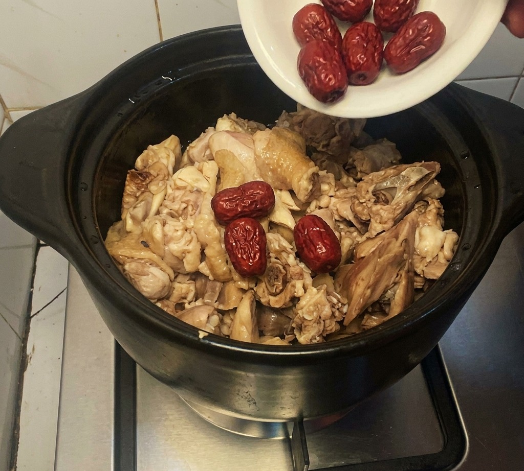 A small white plate pouring whole dried red dates into a clay pot filled with partially cooked chicken pieces.