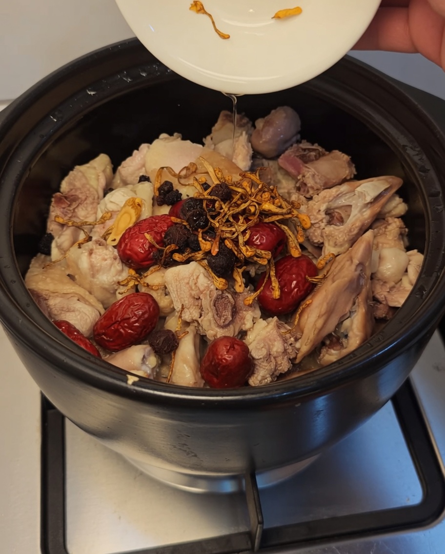 Cordyceps flowers, dark goji berries, and longan being added to the chicken and red dates in a clay pot.