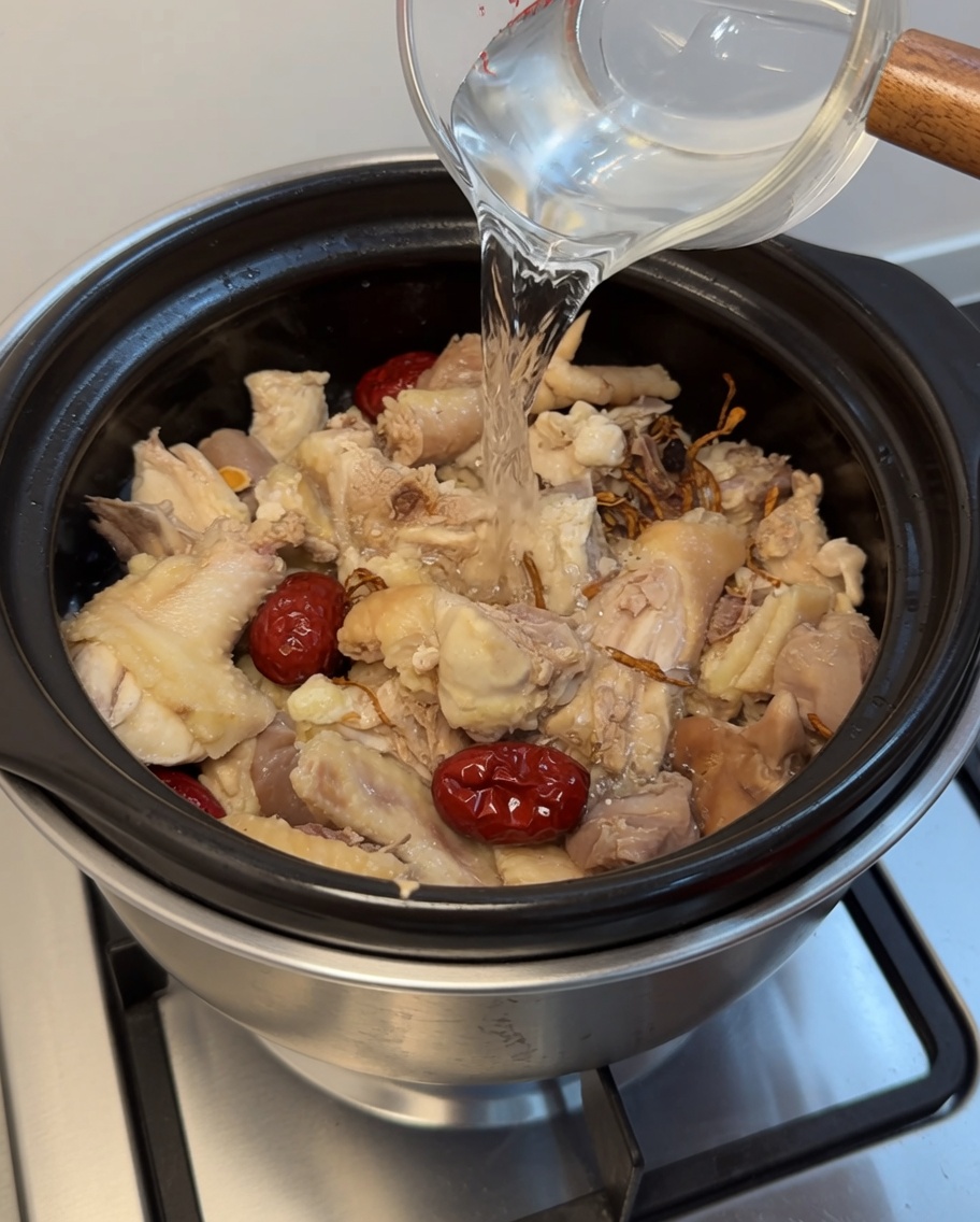Clear water being poured from a glass measuring cup into a clay pot filled with chicken, red dates, and herbs.