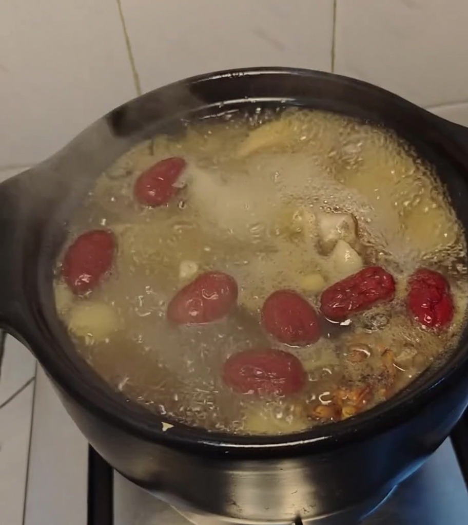 Chicken soup boiling vigorously in a black clay pot with large red dates floating on the surface.