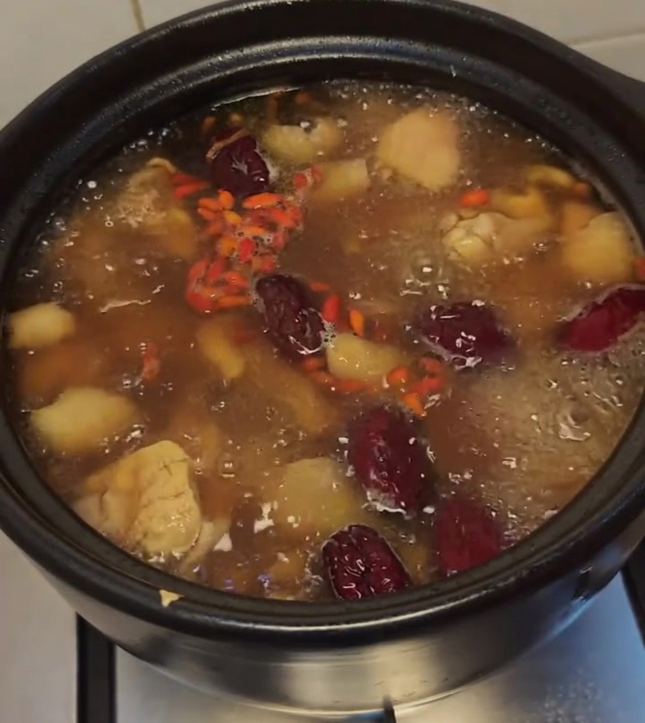 Boiling chicken soup in a black clay pot, garnished with vibrant red goji berries and large red dates.