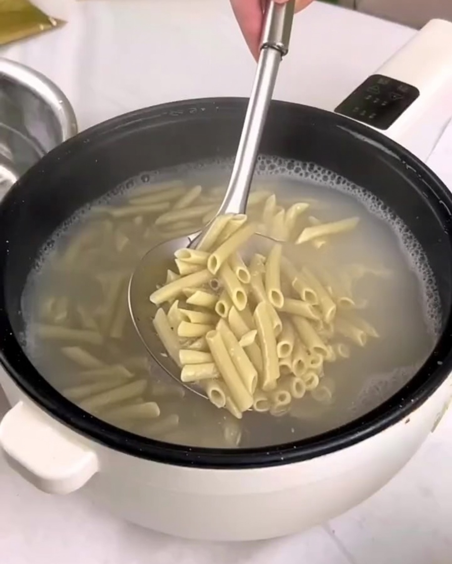 Cooked penne pasta being transferred from a pot into a bowl of cold water using a slotted spoon.