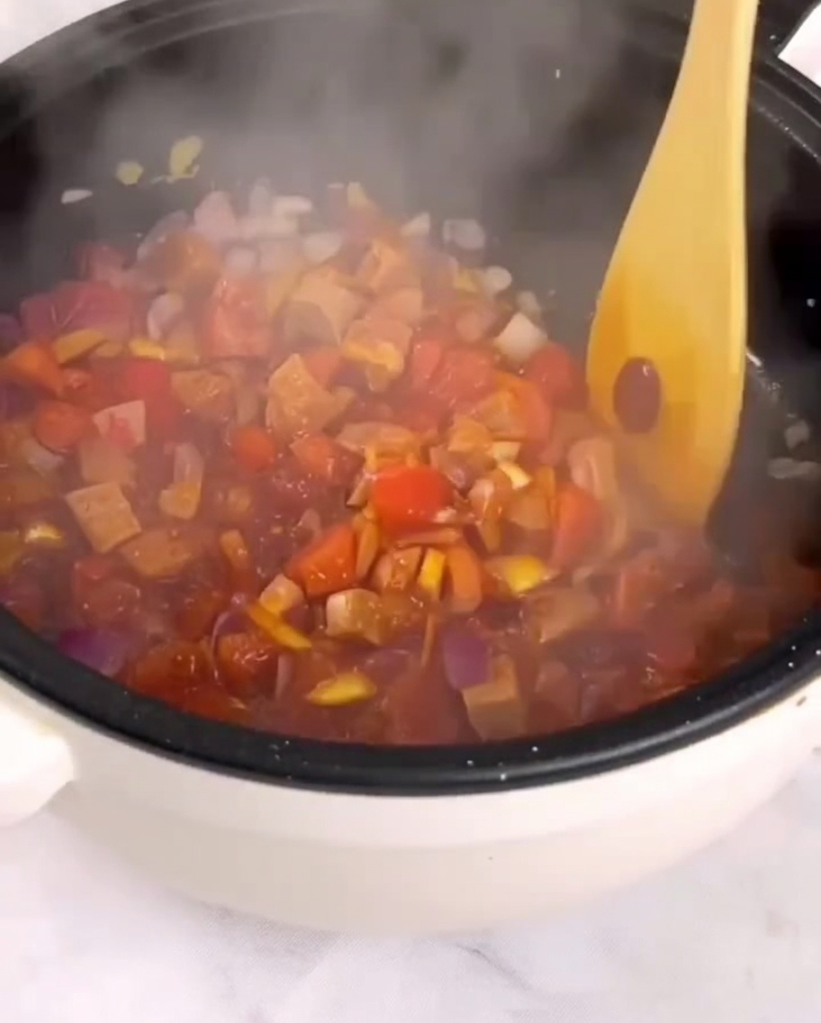 Diced tomatoes, ham, and vegetables being stir-fried in a pan with a wooden spatula.