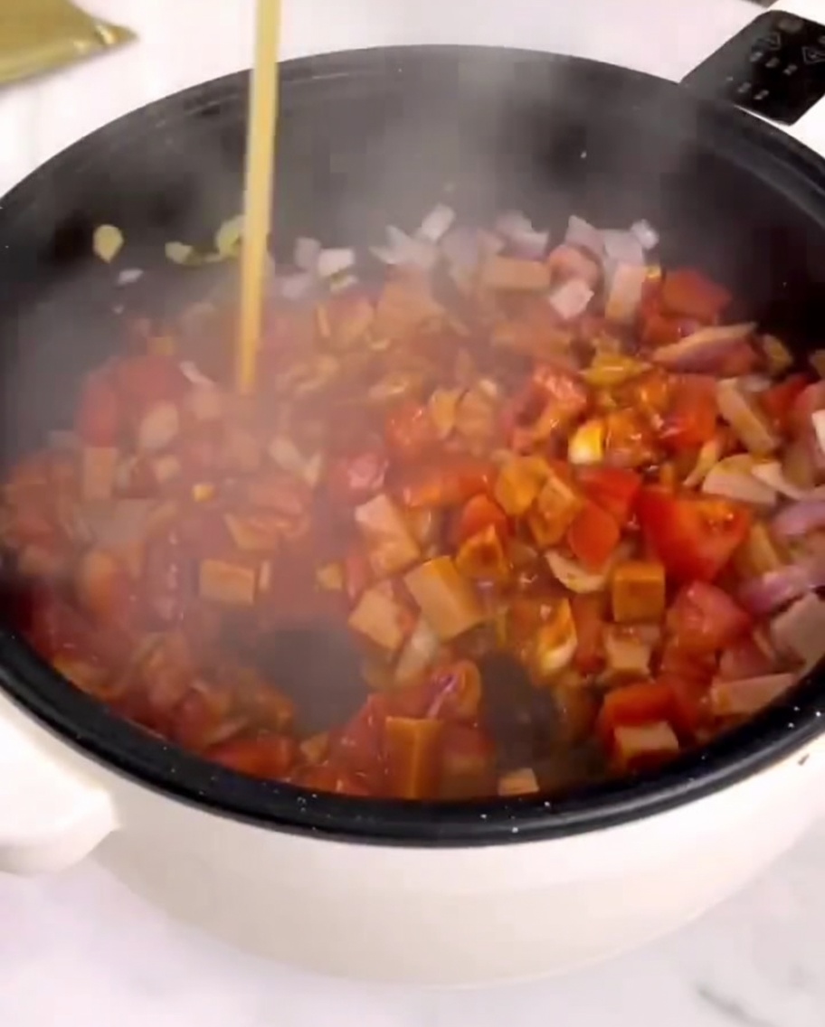Pasta sauce being poured into a pan with stir-fried vegetables and ham.