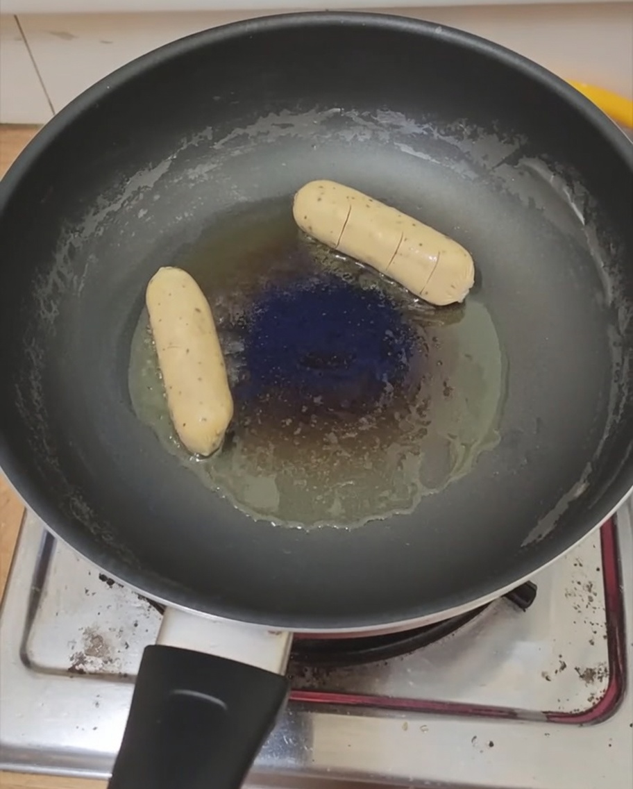 A sunny-side-up egg and a sausage frying in a pan.
