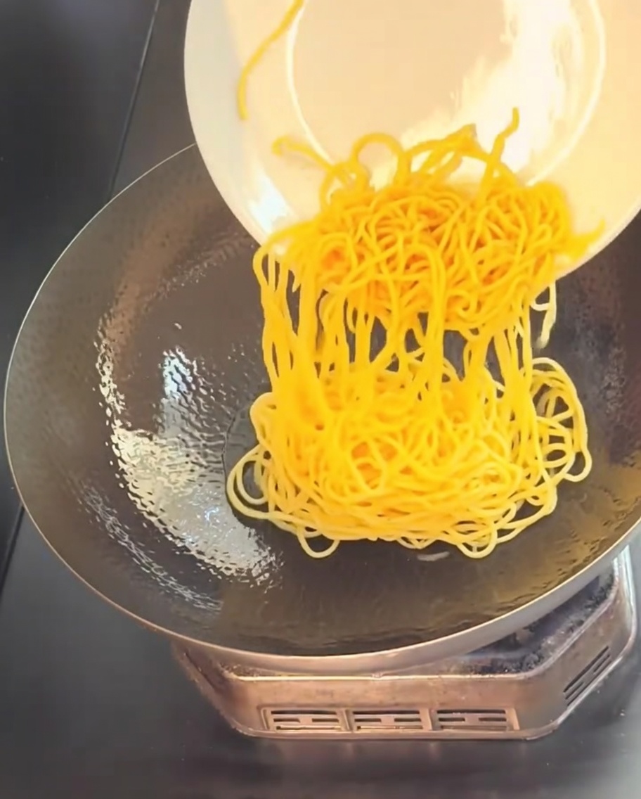 Cooked yellow spaghetti being slid from a white plate into a hot, oiled wok.