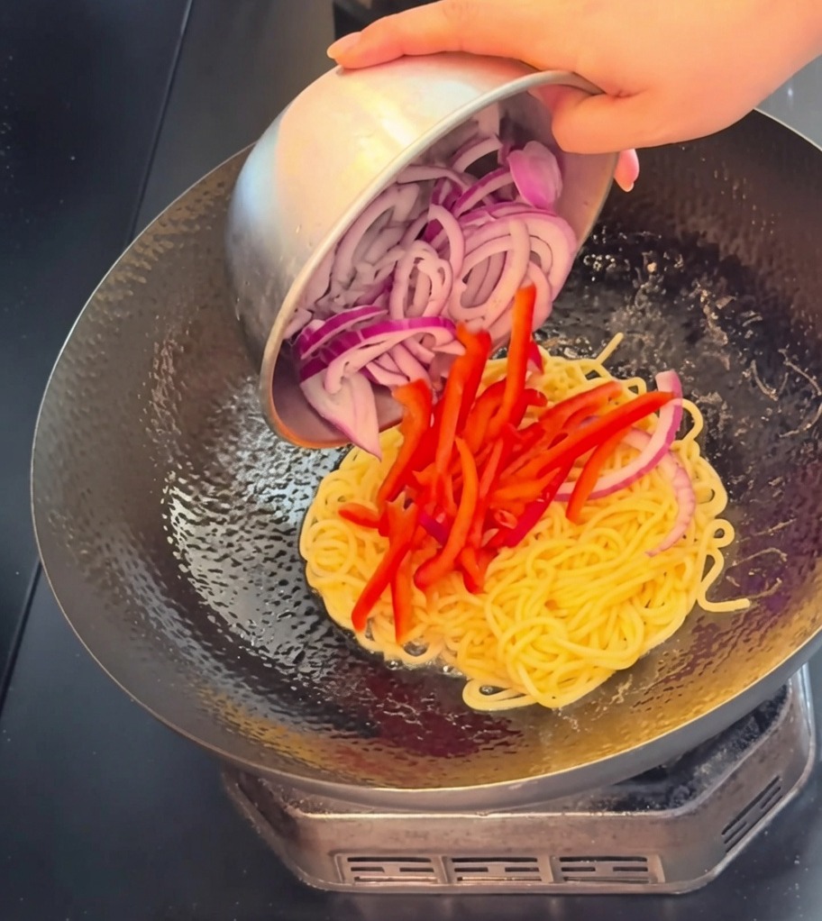 A close-up view of beef slices, vibrant red pepper strips, and purple onion slices being poured into a wok with cooked spaghetti.