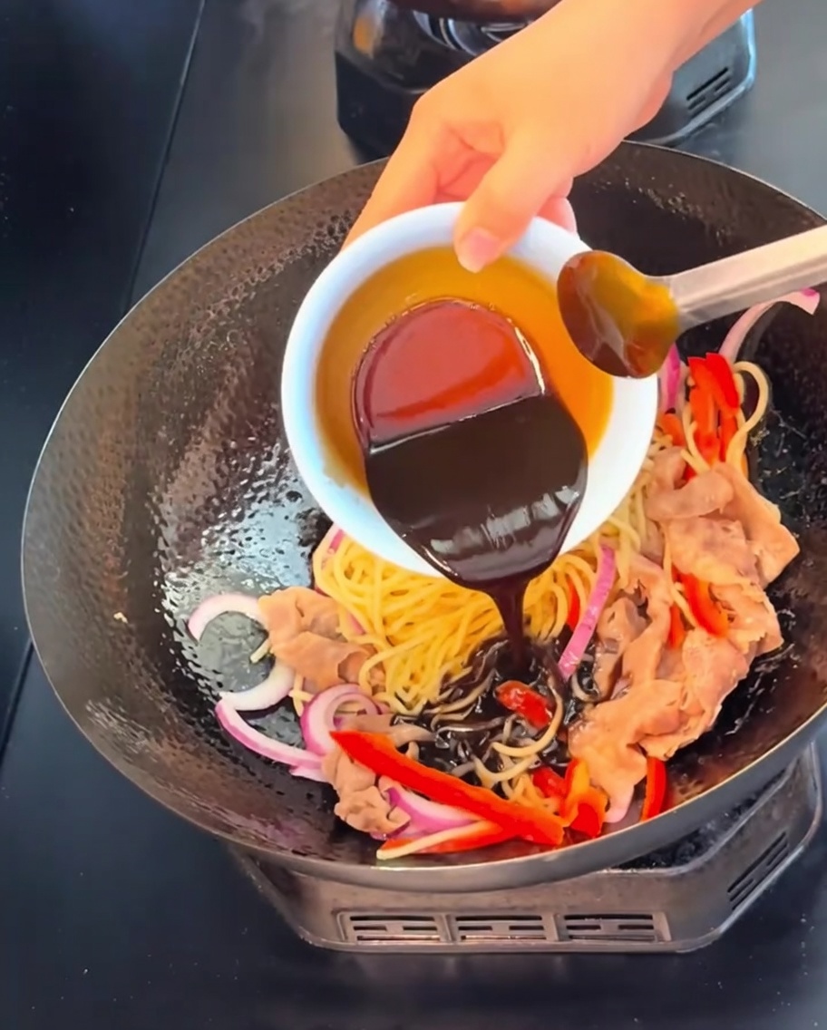 A dark, rich black pepper sauce being poured from a small white bowl over the noodles, beef, and vegetables in a wok.