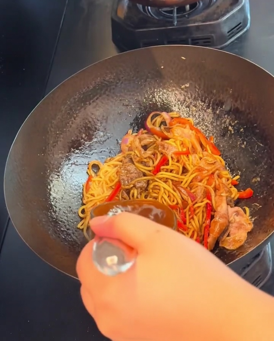 A hand holding a spoon, tossing and stir-frying the black pepper beef spaghetti over high heat in a wok.