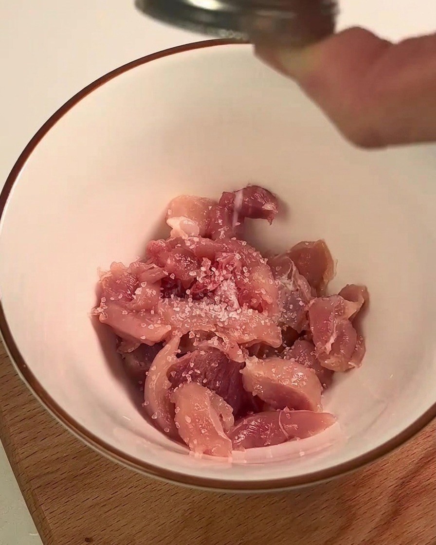 Chicken leg meat being seasoned with salt and lemon juice in a white bowl.