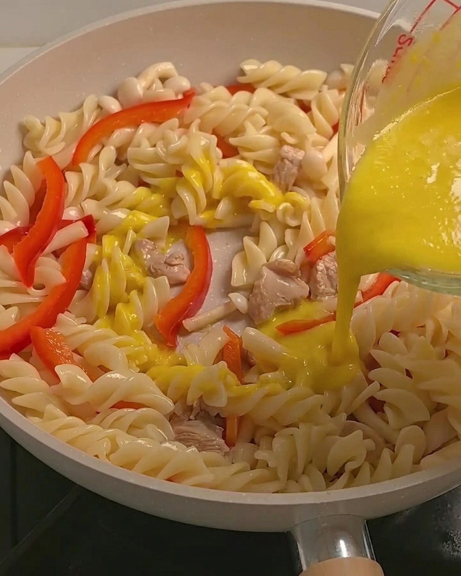 Creamy yellow pumpkin sauce being poured over fusilli pasta, chicken, and vegetables in a pan.