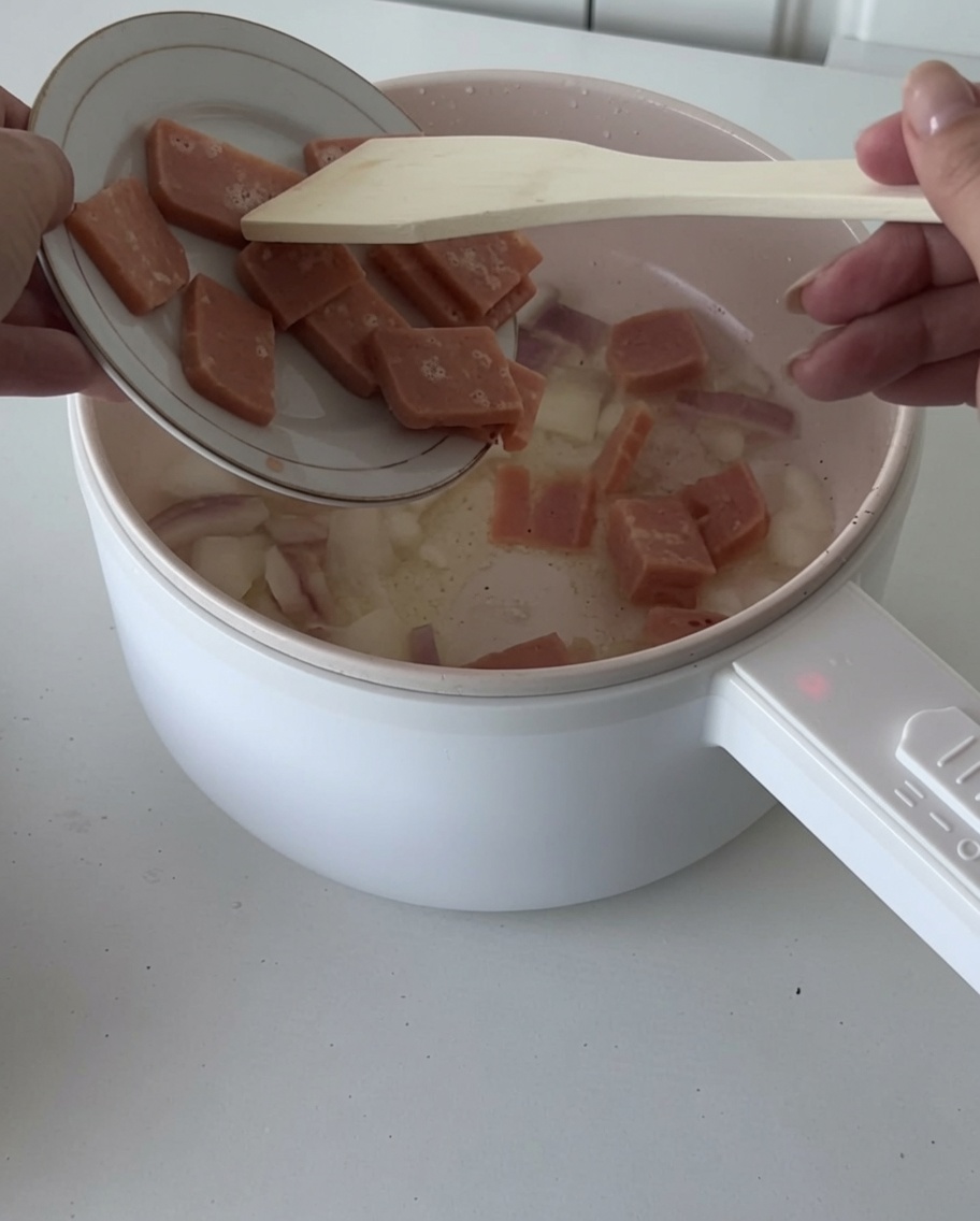 Close-up of bacon pieces being added to a pan with translucent, sautéed onion pieces.