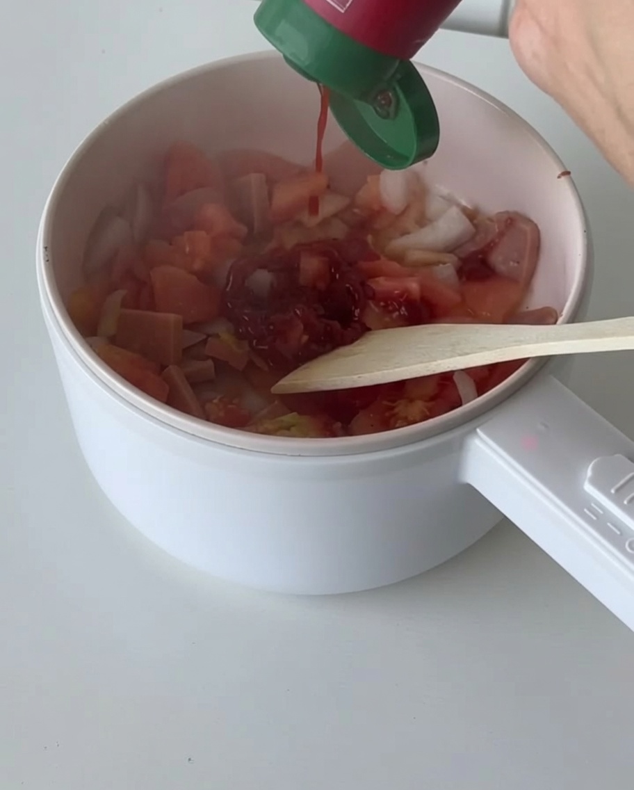 Red tomato sauce being poured from a squeeze bottle into the pan with the vegetable and bacon mixture.