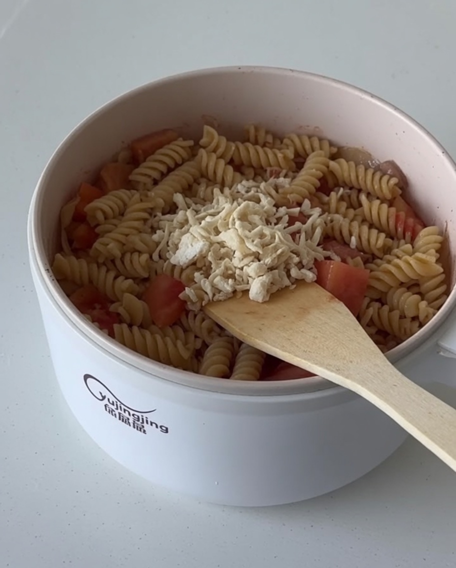Shredded cheese being sprinkled over the finished fusilli pasta mixture in the pan.