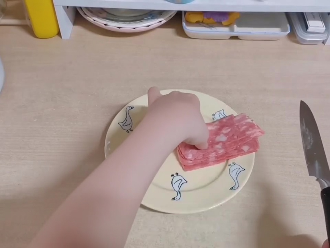 A persons hand using a knife to slice strips of raw bacon into smaller pieces on a yellow plate.