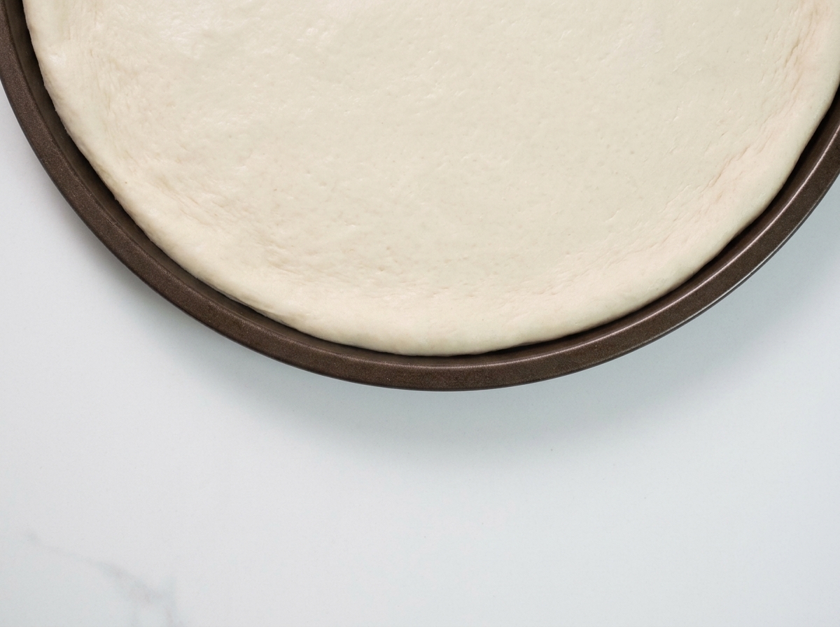 A spoonful of black truffle sauce being spread onto a raw pizza dough base.