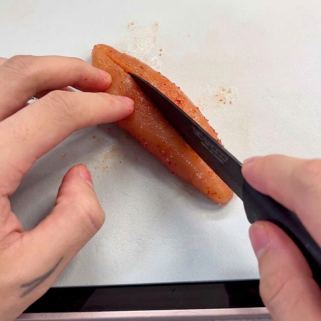 Hands holding a Mentaiko sac and using a knife to scrape out the pink roe on a cutting board.